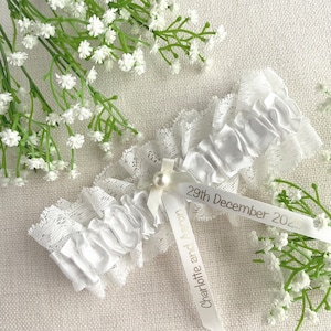 May include: White lace garter with a satin bow and a pearl accent. The garter has a white satin ribbon attached with the text "Charlotte and Aaron 29th December 2023".