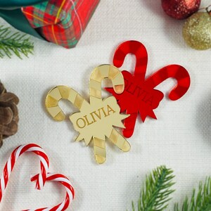 May include: Gold and red Christmas candy cane decorations, one with the name "OLIVIA". The ornaments are on a white surface with festive items, including a wrapped gift and pine needles.