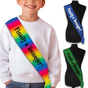 May include: A rainbow sash with the text "Finally a Teenager!" is worn by a person in a white sweatshirt. Two other sashes are shown, one blue and one green, each with the same text.
