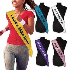 May include: A set of five colourful satin sashes with personalised text. The sashes are yellow, blue, white, purple, and pink. The text on the sashes reads "Laura's 200th Run!", "Jack's 50th Run!", "Matt's 150th Run!", "Oliver's 100th Run!", and "Amy's 15th Run!"