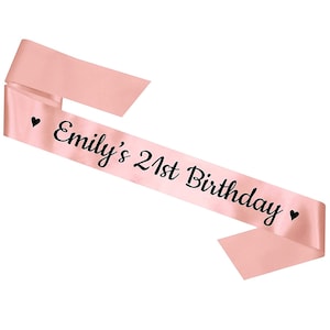 May include: Pink satin sash with black text that reads "Emily's 21st Birthday" with a heart on each side.