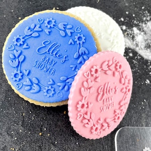 May include: Two round cookies decorated with blue and pink icing. The cookies have a floral design and the text "Ellie's Baby Shower" on them.