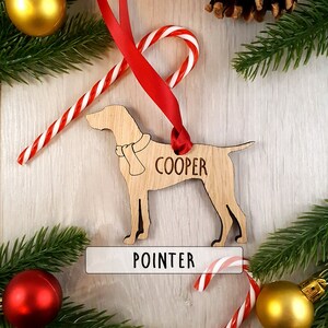 May include: Wooden Christmas ornament shaped like a Pointer dog with the name "COOPER" engraved on it. The ornament is hanging from a red ribbon and has a white label with the word "POINTER" on it.