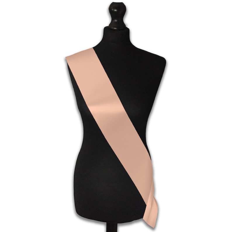 May include: A peach-colored satin sash, displayed on a black mannequin. The sash is angled diagonally across the mannequin's torso.