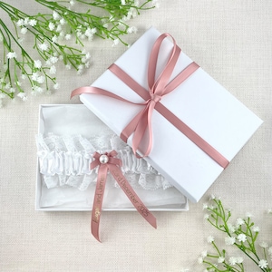 K&ouml;nnte beinhalten: Ein wei&szlig;es Spitzenstrumpfband mit einer Schleife aus Satin und einer Perle. Das Strumpfband befindet sich in einer wei&szlig;en Schachtel mit einem rosa Band und dem Text "Abigail und James 3. Dezember 2025".