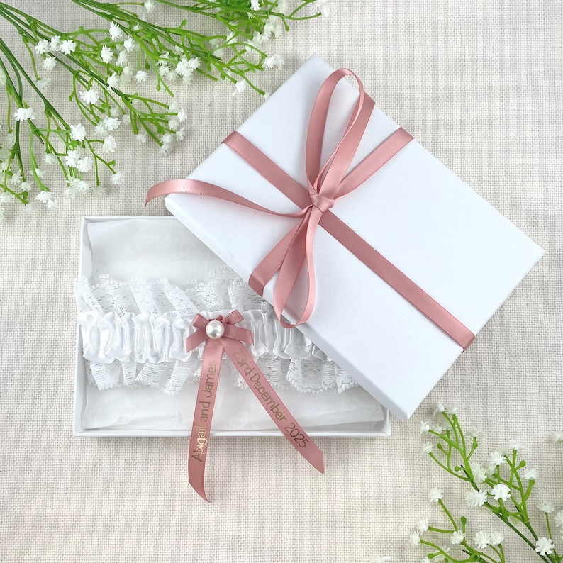 K&ouml;nnte beinhalten: Ein wei&szlig;es Spitzenstrumpfband mit einer Schleife aus Satin und einer Perle. Das Strumpfband befindet sich in einer wei&szlig;en Schachtel mit einem rosa Band und dem Text "Abigail und James 3. Dezember 2025".
