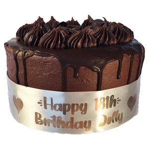 May include: A chocolate cake with chocolate frosting and a chocolate ganache drip. The cake is decorated with a silver ribbon with the text "Happy 18th Birthday Dolly" in gold lettering.