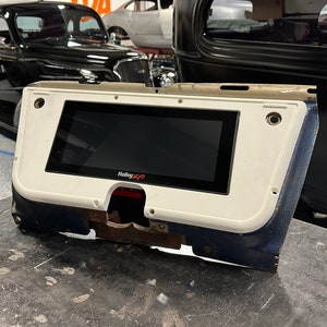 May include: A white and blue car dashboard component with a black screen displaying the Holley EFI logo. The component is rectangular with rounded edges and is set against a garage backdrop with classic cars.