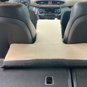 May include: Beige car seat cushion with a black border. The cushion is shaped like a T and is placed between the two front seats of a car.