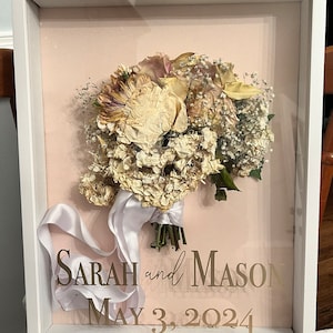 Puede incluir: Un marco blanco con fondo rosa, que muestra un ramo de boda preservado de flores secas. El ramo incluye rosas blancas, hortensias y aliento de bebé. El texto "SARAH and MASON MAY 3, 2024" está impreso en letras doradas sobre el fondo rosa.