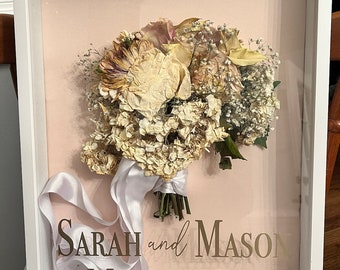 May include: A white frame with a pink background, showcasing a preserved wedding bouquet of dried flowers. The bouquet includes white roses, hydrangeas, and baby's breath. The text "SARAH and MASON MAY 3, 2024" is printed in gold lettering on the pink background.