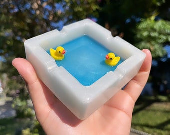 May include: A square, white ashtray with a light blue interior, featuring two small yellow rubber duckies floating inside. The ashtray is held in a hand, with a blurred outdoor background.