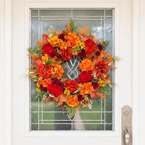 May include: A circular autumn wreath featuring a dense arrangement of artificial flowers and foliage in vibrant shades of deep red, burnt orange, and golden yellow, accented with green leaves, displayed on a white door with a glass panel insert.