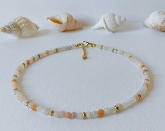 May include: Delicate beaded necklace featuring small, round, peach, white, and gray beads. Gold-colored spacers and a gold-colored clasp complete the necklace. The necklace is displayed on a white surface with seashells in the background.
