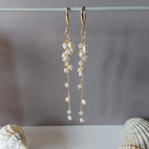 May include: Elegant gold-tone earrings with cascading white pearls. The earrings feature a cluster of pearls at the top, with delicate chains of smaller pearls dangling below. The design is simple yet sophisticated, perfect for special occasions.