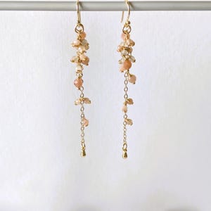 Könnte beinhalten: Hängeohrringe mit pfirsichfarbenen Perlen und goldfarbenen Metallakzenten. Die Ohrringe haben oben eine Ansammlung von Perlen, mit einer Kette und weiteren Perlen, die zu einem kleinen Tropfenanhänger herabfallen. Weißer Hintergrund.