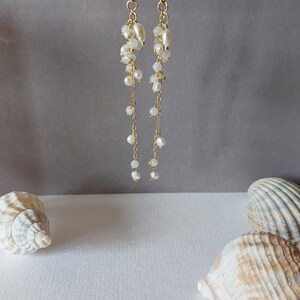 May include: Elegant gold-tone earrings with cascading white pearls and smaller beads. The earrings hang from a gold hook and are displayed against a neutral gray background. Seashells are in the foreground, suggesting a beach theme.