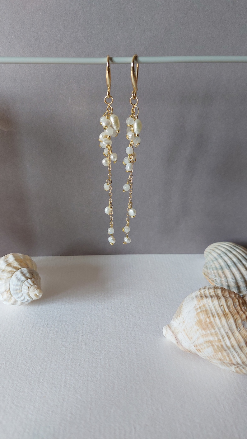 May include: Elegant gold-tone earrings with cascading white pearls and smaller beads. The earrings hang from a gold hook and are displayed against a neutral gray background. Seashells are in the foreground, suggesting a beach theme.