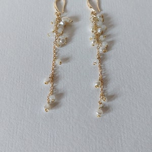May include: A pair of gold-toned drop earrings. Each earring features a gold hook, a cluster of small white beads, and a delicate gold chain with dangling white pearls. The earrings are displayed on a white surface.