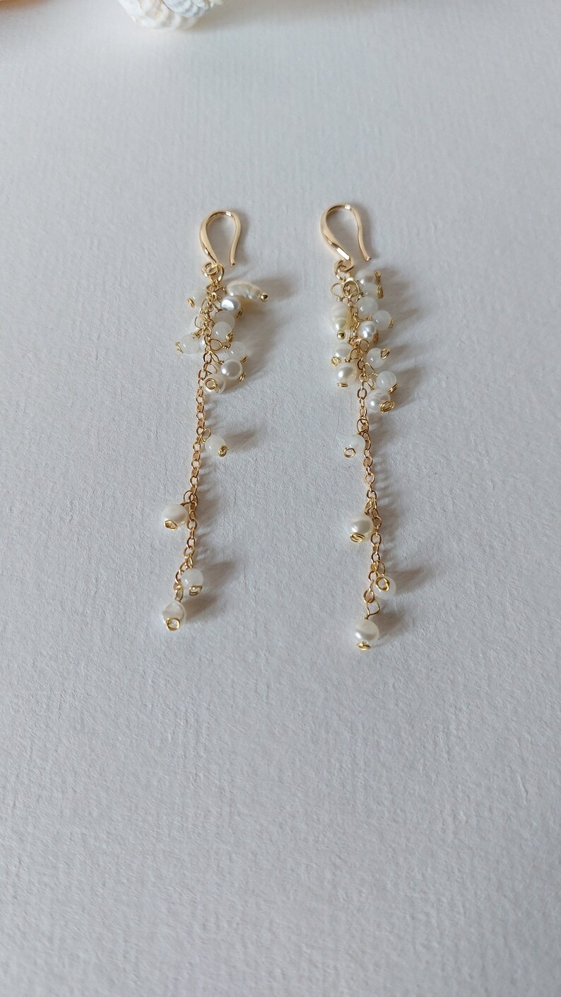 May include: A pair of gold-toned drop earrings. Each earring features a gold hook, a cluster of small white beads, and a delicate gold chain with dangling white pearls. The earrings are displayed on a white surface.