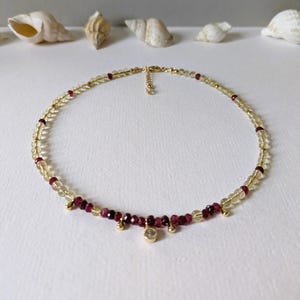 Può includere: Delicata collana con piccole pietre preziose giallo chiaro e rosso intenso. La collana ha dettagli color oro e un piccolo pendente trasparente. È esposta su una superficie bianca con conchiglie sullo sfondo.