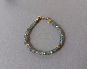 May include: A delicate bracelet featuring small, faceted, gray-green beads with flashes of blue. The bracelet is accented with gold-colored square beads and a gold-colored clasp. The background includes seashells on a gray surface.