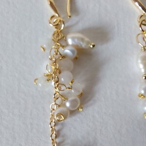 May include: Gold-tone earrings with a cluster of white pearls and small, round, white beads. The earrings have a delicate chain and a curved hook closure. The design is elegant and suitable for various occasions.