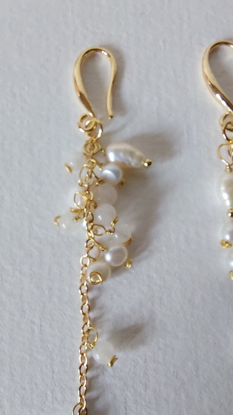 May include: Gold-tone earrings with a cluster of white pearls and small, round, white beads. The earrings have a delicate chain and a curved hook closure. The design is elegant and suitable for various occasions.