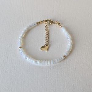 May include: Delicate white beaded bracelet with gold-colored accents. The bracelet features small, round white beads and gold-colored beads. A small gold heart charm hangs from the adjustable chain clasp.