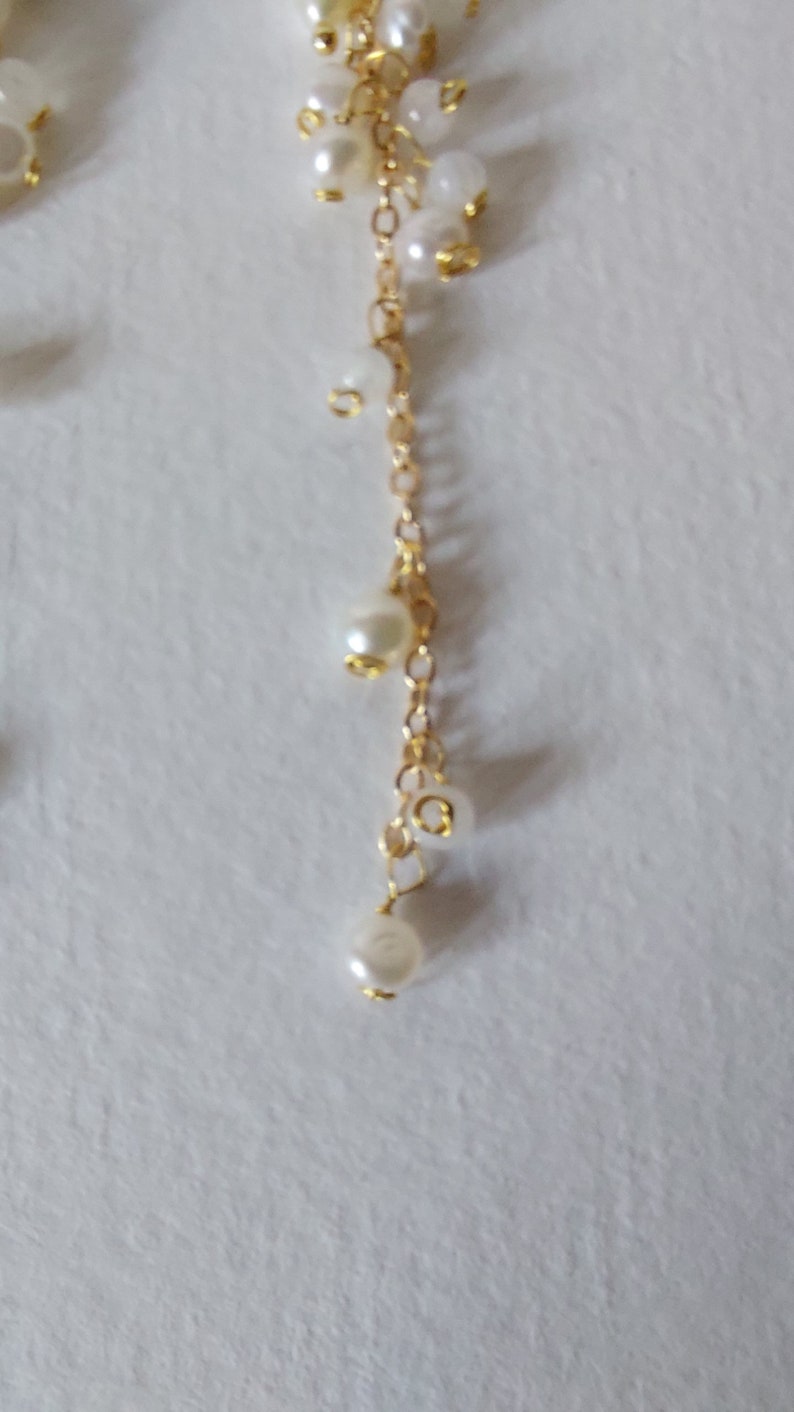May include: A delicate gold chain adorned with clusters of small, creamy white pearls. The chain cascades downward, with larger pearls suspended at intervals. The pearls have a subtle luster, creating an elegant and timeless aesthetic.