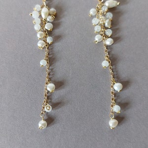 May include: A pair of elegant gold-tone earrings with a cascading design. Each earring features a cluster of small, white pearls at the top, with a delicate chain of gold links and larger pearls that dangle below. The earrings have a hook closure.