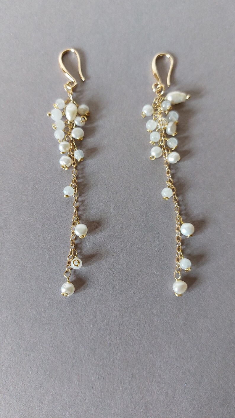 May include: A pair of elegant gold-tone earrings with a cascading design. Each earring features a cluster of small, white pearls at the top, with a delicate chain of gold links and larger pearls that dangle below. The earrings have a hook closure.