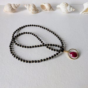 Op de afbeelding: Een zwarte kralenketting met een gouden hanger met een grote, ovaalvormige, rode edelsteen. De ketting wordt gepresenteerd op een wit oppervlak, met schelpen op de achtergrond.