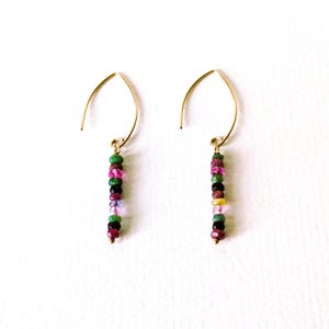 Puede incluir: Un par de pendientes de aro dorados. Cada pendiente presenta una hebra vertical de piedras preciosas redondas, pequeñas y coloridas en tonos verdes, rosas, morados y amarillos. Los pendientes se muestran sobre un fondo blanco.