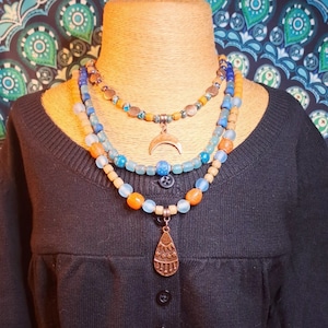 Puede incluir: Collar de capas con cuentas azules, naranjas y de madera. El collar presenta un dije de media luna y un dije en forma de lágrima con un diseño geométrico.