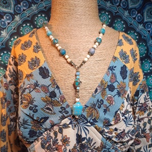 Pode incluir: Um colar de contas turquesa e branco com um pingente que apresenta um amuleto com padrão turquesa e branco. O colar é exibido em um manequim que usa uma blusa com estampa floral azul e amarela.