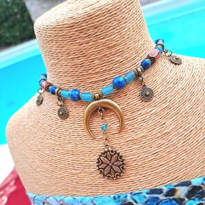 Puede incluir: Un collar gargantilla de cuentas azules y marrones con un dije de media luna y un dije redondo con un diseño floral.