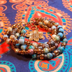 Op de afbeelding: Meerstrengige armband met turquoise, bruine en witte kralen, en een gouden bloembedel.