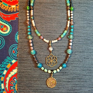 Pode incluir: Um colar de várias camadas com contas pretas, brancas, cor-de-rosa e azuis. O colar tem dois pingentes, um com um desenho de flor da vida e o outro com uma lua crescente e estrelas.