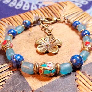 Op de afbeelding: Een kralenarmband met blauwe, turquoise en rode accenten. De armband heeft blauwe lapis lazuli kralen, turquoise glazen kralen en decoratieve metalen kralen. Een bronzen bloemvormige bedel hangt aan de sluiting.