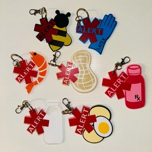 Può includere: Una collezione di portachiavi colorati per l'allerta allergie. I disegni includono un'ape, gamberetti, arachidi, bottiglia di latte, uovo e un flacone di pillole rosa. Ogni portachiavi ha una croce rossa con la scritta "ALERT".