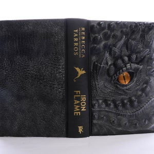 May include: A black hardcover book with a dragon embossed on the cover. The spine of the book is black with gold lettering that reads "Iron Flame" by Rebecca Yarros.