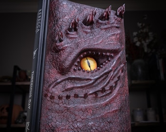 May include: A hardcover book titled Rebecca Yarros Threshing Day with a dark red, textured dragon scale cover featuring a prominent, realistic yellow eye in the center and several raised, sharp, dark red spikes along the top edge.
