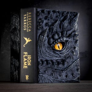 May include: A black book with a dragon eye embossed on the cover. The book title is "Iron Flame" by Rebecca Yarros.