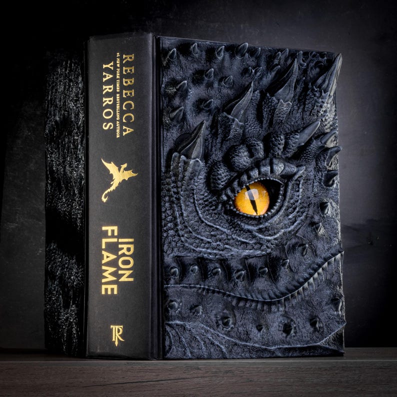 May include: A black book with a dragon eye embossed on the cover. The book title is "Iron Flame" by Rebecca Yarros.