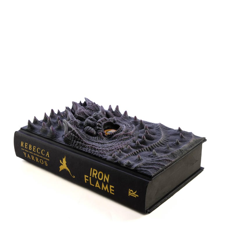 May include: A black hardcover book with a dragon embossed on the cover. The dragon's eye is a yellow gemstone. The title of the book is "Iron Flame" and the author is "Rebecca Yarros".