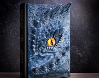 May include: A blue leather-bound book with a dragon's eye embossed on the cover. The eye is yellow with a black pupil. The book is titled "Rebecca Onyx".
