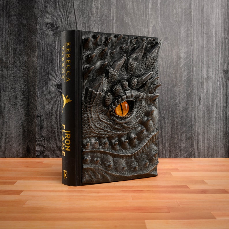 May include: A black hardcover book with a dragon embossed on the cover. The dragon has a yellow eye and a textured, scaly design. The book title is "Iron Flame" by Rebecca Yarros.