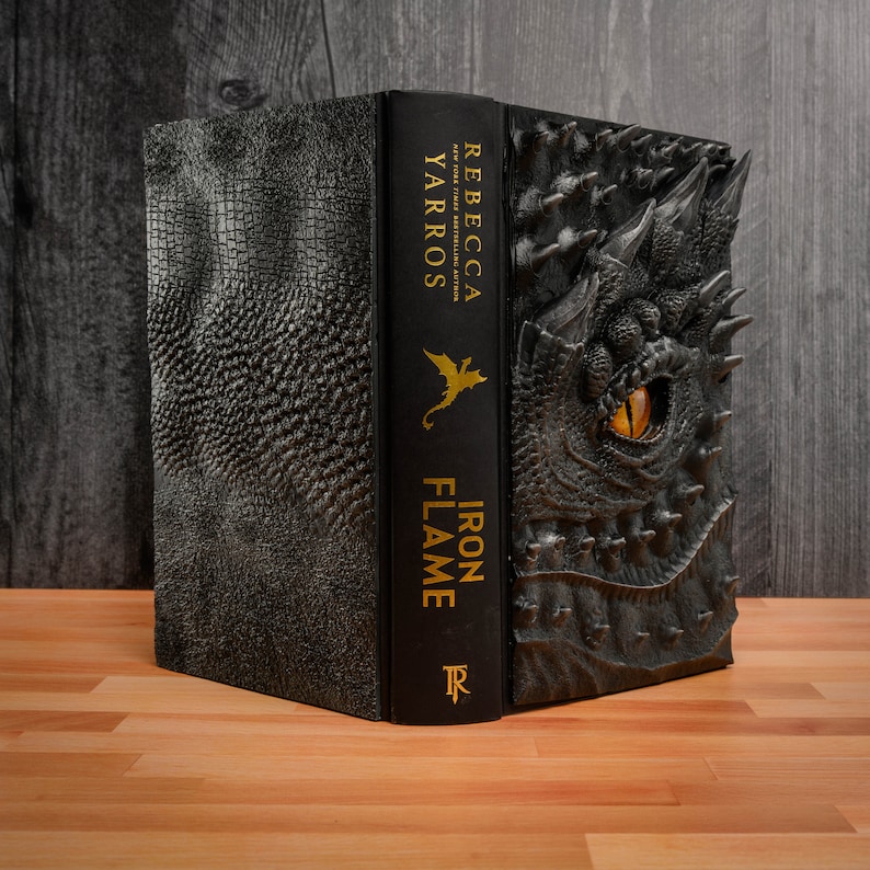 May include: A black hardcover book with a dragon embossed on the cover. The title "Iron Flame" and the author "Rebecca Yarros" are printed in gold on the spine.