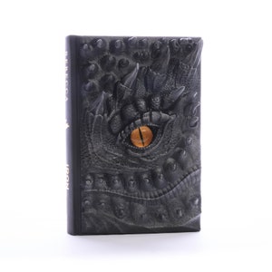 May include: Black leather-bound book with a dragon eye embossed on the cover. The eye is yellow with a black pupil. The book has the words "Rebecca Iron" printed on the spine.